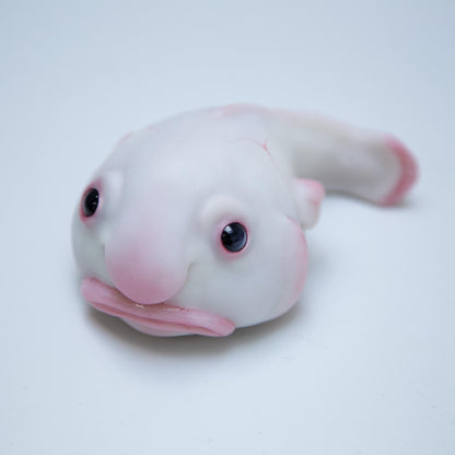 A small sculpture of a blob fish. It has white skin, a large head, a wide sad but slightly stupid face. The sculpture is made of polymer and epoxy