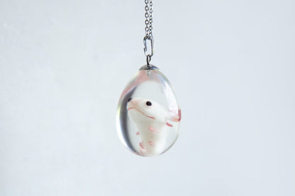 a small axolotl embryo figurine with black eyes, placed inside a crystal clear egg-shaped pendant made of epoxy resin