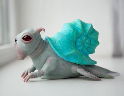A fantasy creature, an alien character living in a shell. It has fins with transparent membranes, large eyes and antennae on its head. Its house on its back is a blue curl shell. The sculpture is made of gray-blue and blue polymer