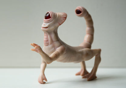 Alien monster cyclops, a strange and creepy creature from other worlds. A hybrid of a cyclops and a grasshopper. The sculpture is beige, the back legs are three-fingered, there are hands in front, and a face with one eye. There is a movable tail with an eye at the end at the back.