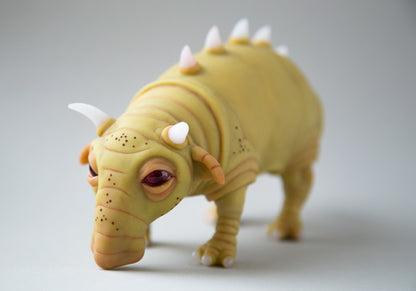 A fantasy alien creature, Make-a-boar. A hybrid of a pig and a bull, the creature has horns, rough skin like a rhinoceros, spikes on its back and legs with three hooves. The sculpture is made of mustard-colored polymer, the horns and hooves are made of white translucent. Created and invented character from Planet Cocoon