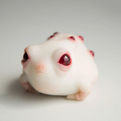 A frog figurine, our interpretation of the African rain frog. It has plump sides, a sad face and a cute butt. It also has red epoxy warts on its back, and large, red eyes. The body is made of white polymer, the sculpture's accents are painted red.