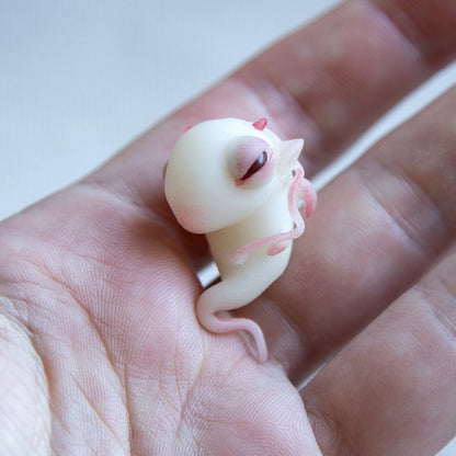 A small dragon embryo, translucent white with red eyes, still has horns, ears and an umbilical cord on its body