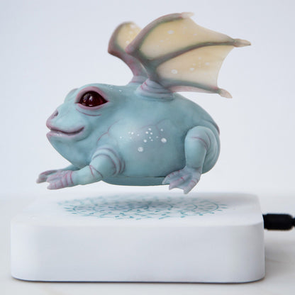 A gray-blue frog figurine with two dragon wings, levitating in an electromagnetic field. Sculptural composition of a winged frog made of polymer, also containing a permanent magnet, and a white electromagnetic stand, levitating sculpture
