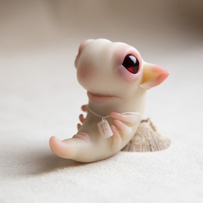 A small figurine of a dragon embryo. It is made of white polymer, has red eyes and a smiling face and six small paws. In addition, it has a necklace with a rune that protects it.