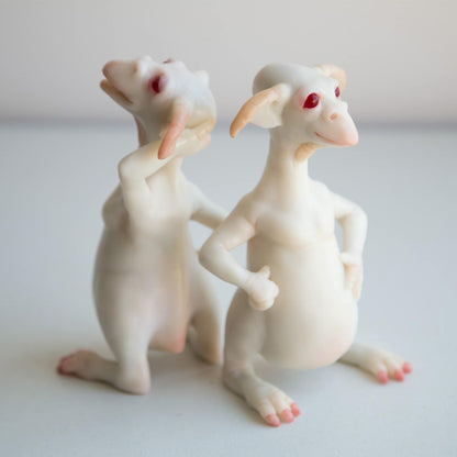 A pair of cheerful goblin twins. They have long necks, big ears, a semi-circular jaw like a toad and legs like a kangaroo. They are albinos with red eyes. The sculptures are made of white polymer.