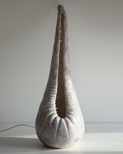 A sculptural ceramic table lamp shaped like a teardrop or seashell, featuring a ribbed textured surface in a light gray and off-white glaze, shown on a plain light gray background with a power cord visible on the left.