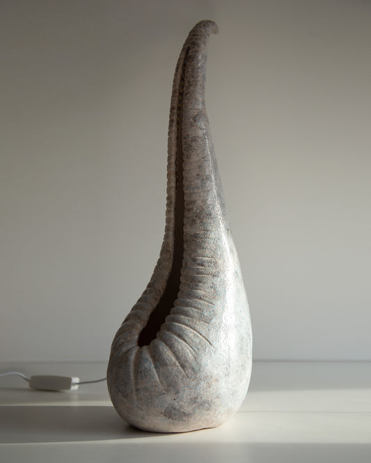 A sculptural teardrop-shaped lamp or sculpture with a textured, metallic silver-gray finish and a long curved neck, standing on a white surface against a neutral gray background.
