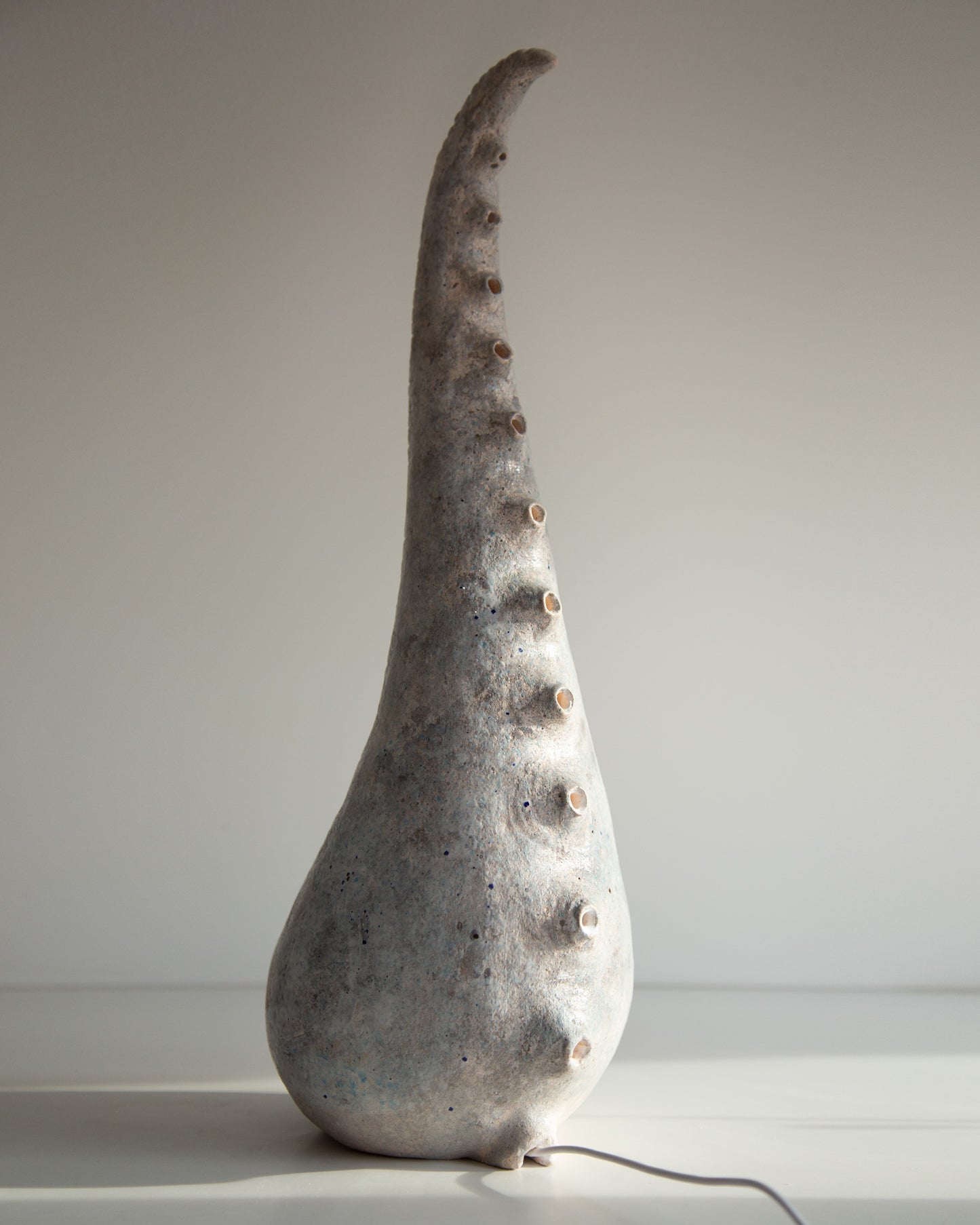 A sculptural ceramic table lamp shaped like a tall, curved shell or horn, with a textured gray and white glaze and small raised bumps along the side, shown on a plain light background.