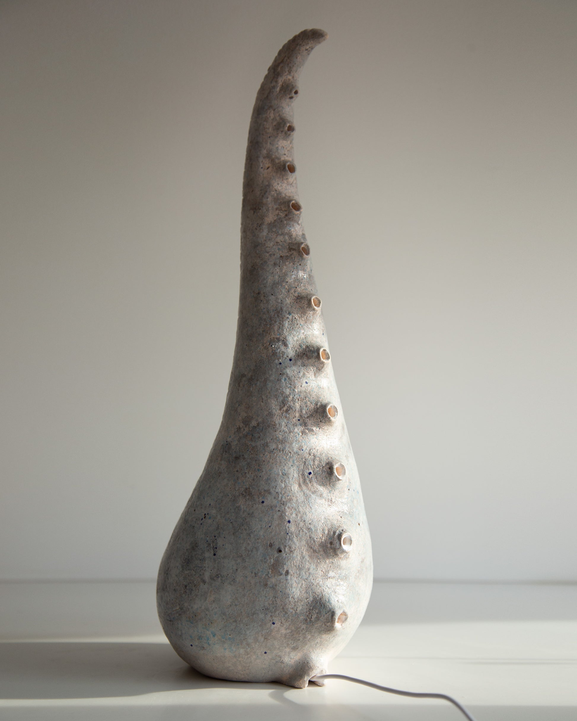 A sculptural ceramic table lamp shaped like a tall, curved shell or horn, with a textured gray and white glaze and small raised bumps along the side, shown on a plain light background.