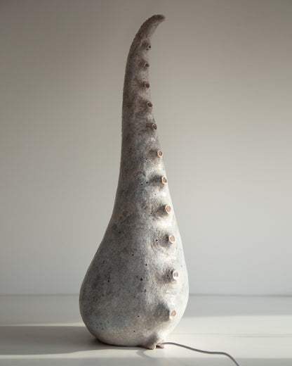 A sculptural ceramic table lamp shaped like a tall, curved shell or horn, with a textured gray and white glaze and small raised bumps along the side, shown on a plain light background.