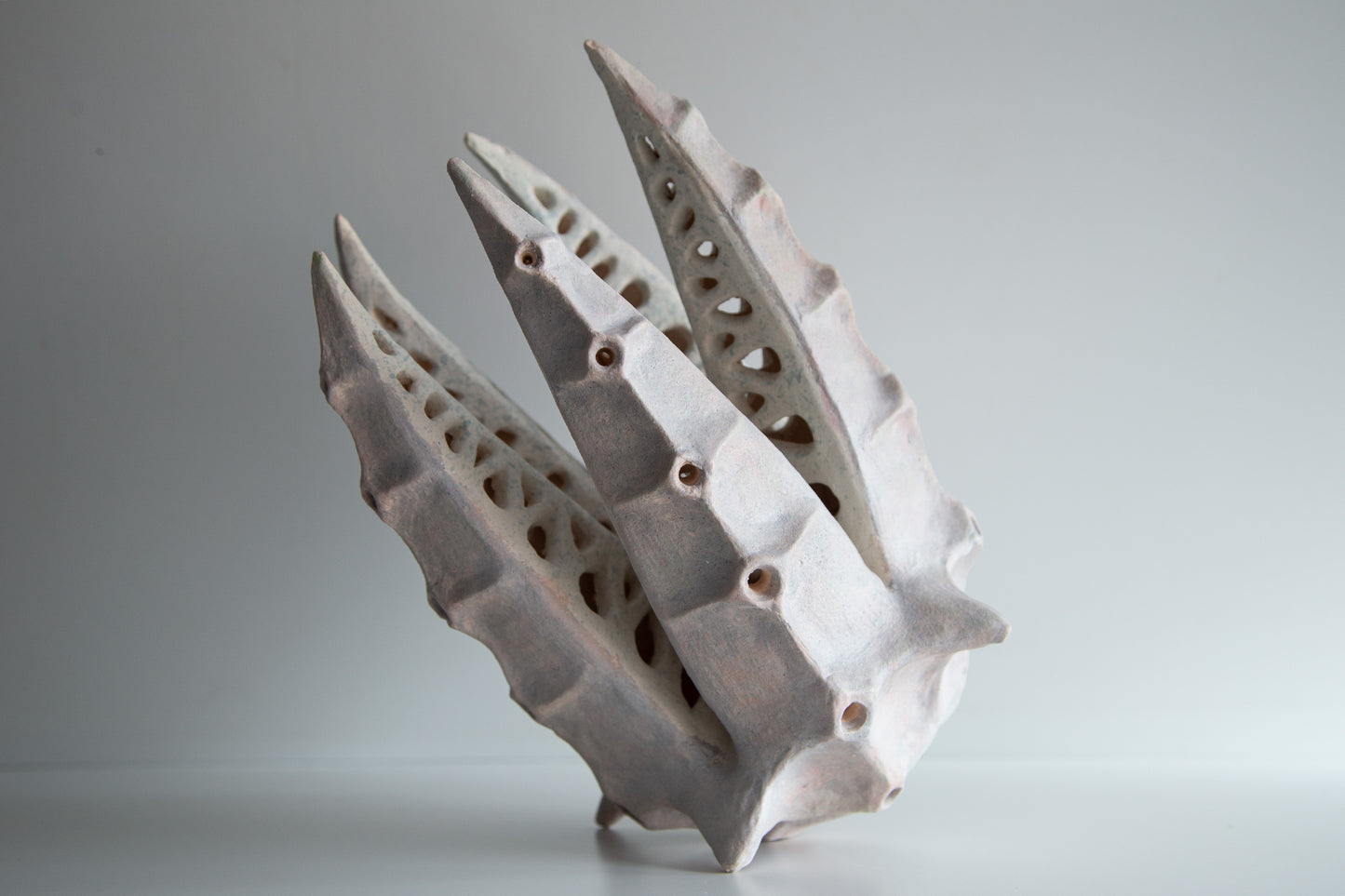 An abstract sculptural ceramic object with three tall, pointed, perforated shell-like forms, photographed on a light gray studio background.