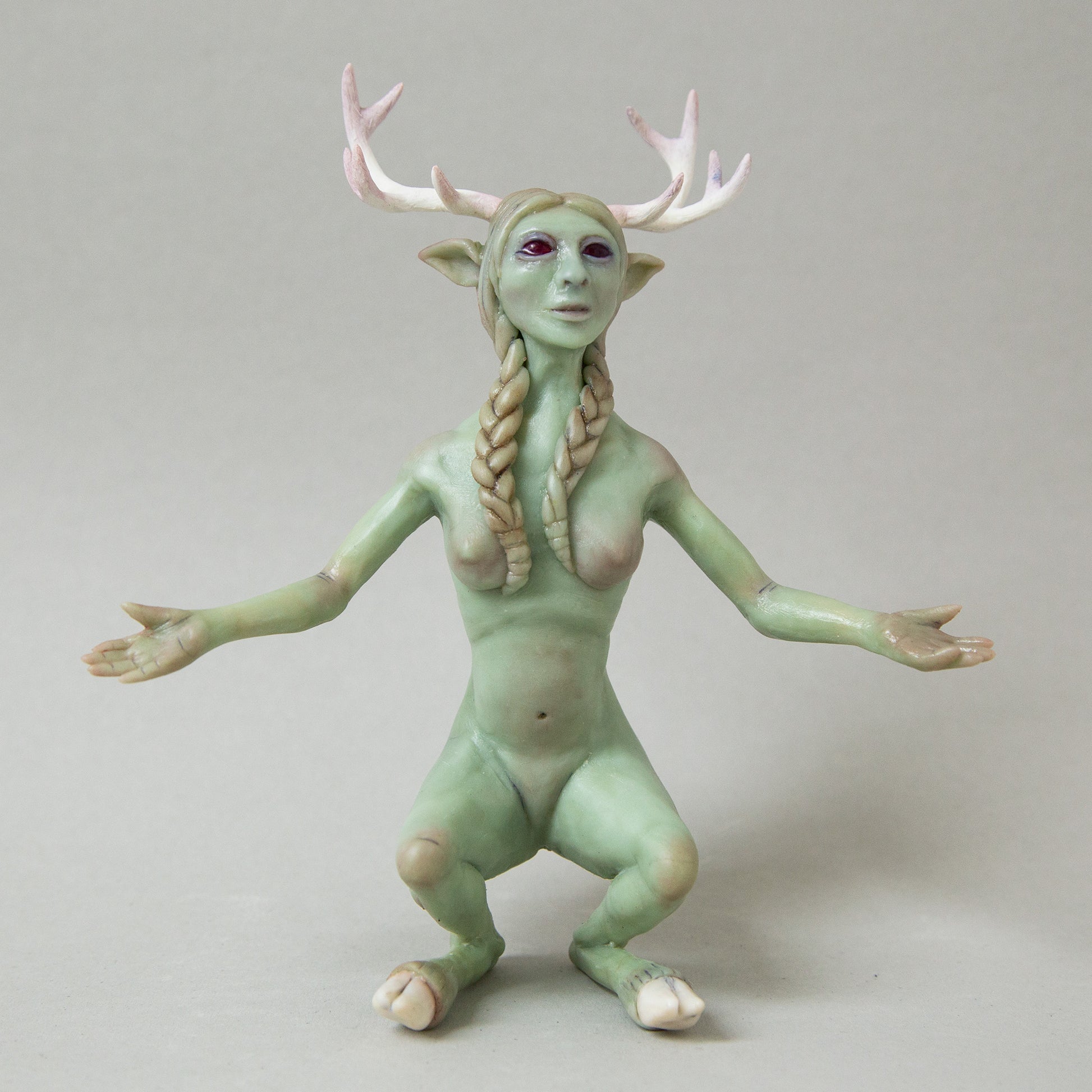 Handcrafted figurine of a mystical humanoid creature with green skin, delicate antlers, pointed ears, and long braided hair. The figure has an otherworldly appearance, blending human and animal features, with open arms and a serene expression.