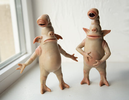 Sculpture of two goblin twins with one eye, goblins-cyclops. One stands in a contented pose with his hands on his stomach, smiling. The second has a confused look, his hands are spread out, and a pose of disappointment or as if he lost something. The sculptures are made of beige polymer, the eyes are made of epoxy