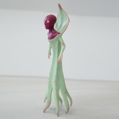 An alien creature from West Virginia folklore, the Flatwoods Monster. It is a humanoid, with red skin and glowing eyes, it also has thin arms with long fingers, its body is wrapped in a green dress with folds. The sculpture is made of green and red polymer, the eyes are made of luminescent epoxy.