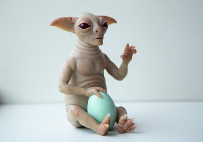 A fantasy character, an alien from the Yoda race. A cute, Yoda-like creature, in a sitting pose with a blue egg, which he holds with his legs and one hand. The other hand is raised up, the index finger is extended upward. The character has beige skin, large goblin ears and a wide face.