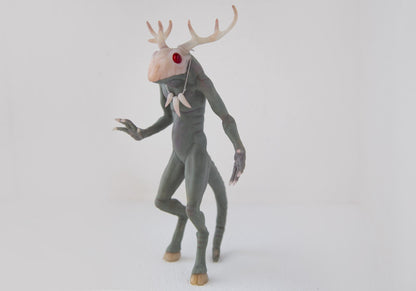 Wendigo, a creepy demon from Minnesota folklore. It has gray skin, elk-like hooved feet, deer antlers on its head, and a skull mask on its face. It has strong arms with long claws, and a long demonic tail at the back. The sculpture is made of gray polymer, the mask, horns, hooves, and claws are translucent. The red eyes are made of epoxy.
