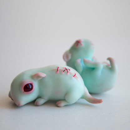 A funny little mouse in the anime style, blue in color, with a stitched scar on his back. He has red eyes and a sad look. The sculpture is made of blue or turquoise polymer, fits in the hand