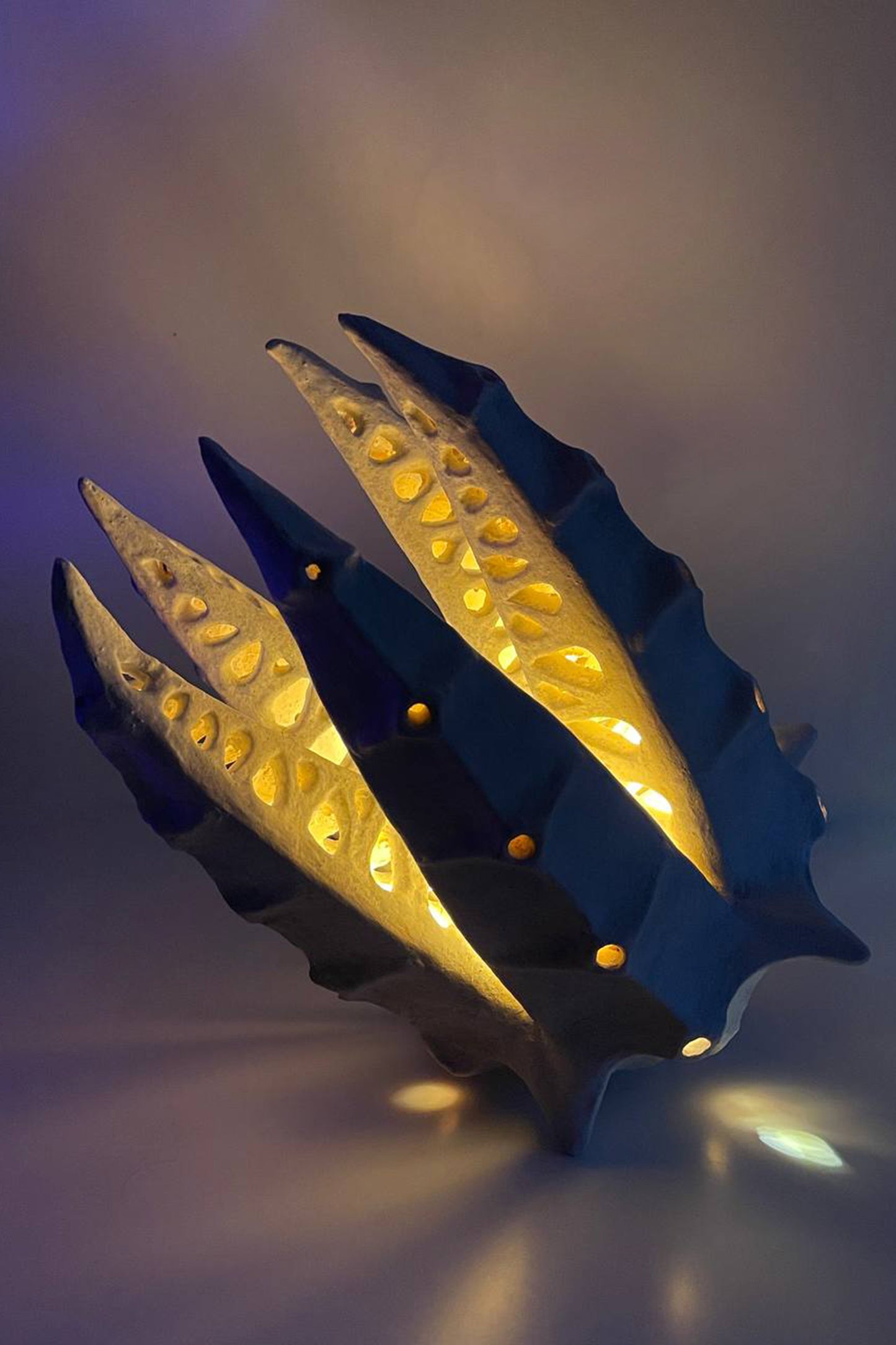 A sculptural modern table lamp with a dark blue and gold leaf-like organic frame, illuminated from within to cast a warm golden glow.