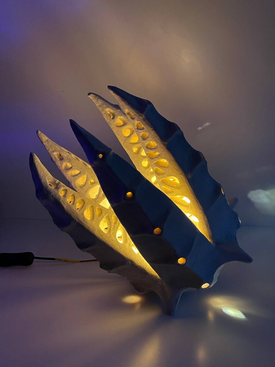 A sculptural modern lamp with a dark blue-gray shell-like form and warm yellow internal lighting, shown on a plain surface with a visible power cord.
