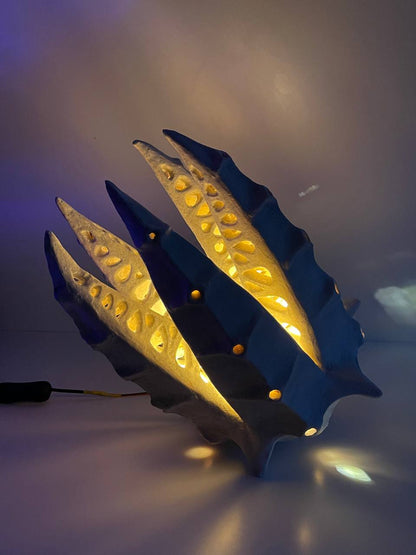 A sculptural modern lamp with a dark blue-gray shell-like form and warm yellow internal lighting, shown on a plain surface with a visible power cord.